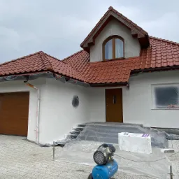 Dom jednorodzinny z nową, czerwoną dachówką i świeżo pomalowaną na biało elewacją, zabezpieczony folią malarską, z widocznym kompresorem i materiałami budowlanymi na podjeździe z kostki.