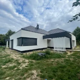 Nowoczesny dom jednorodzinny z białą elewacją, czarnymi akcentami i żaluzjami zewnętrznymi, otoczony trawnikiem i zielenią, widok na pochmurne niebo.