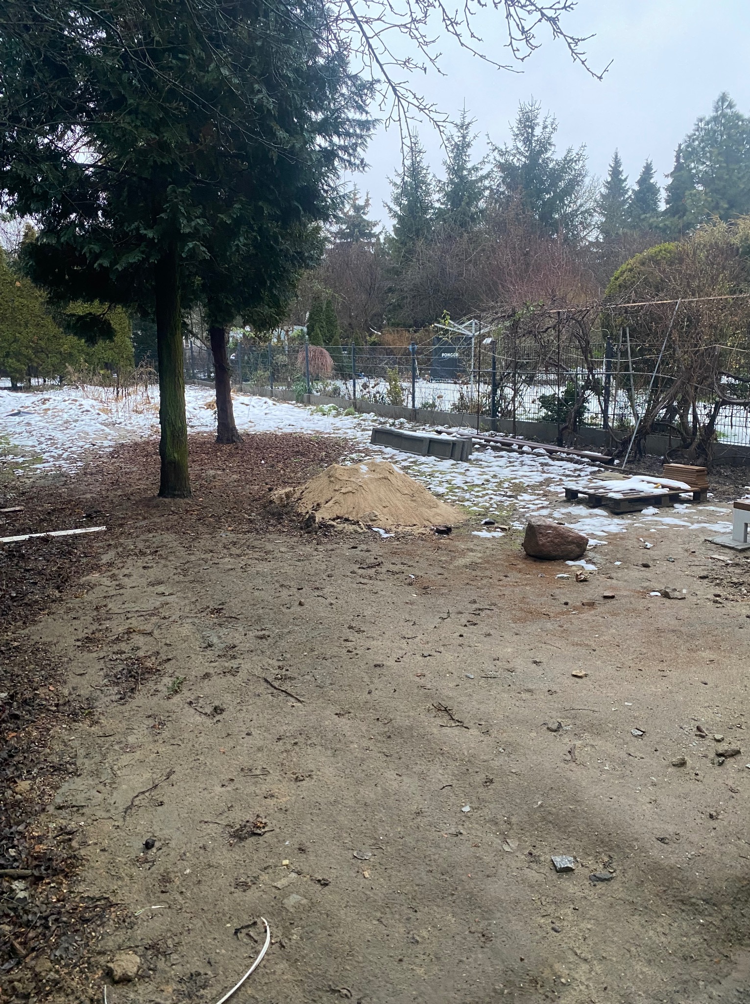 Przygotowanie terenu pod budowę ogrodzenia: sterta piasku, elementy betonowe i drewniane deski na ziemi z resztkami śniegu, drzewa w tle.