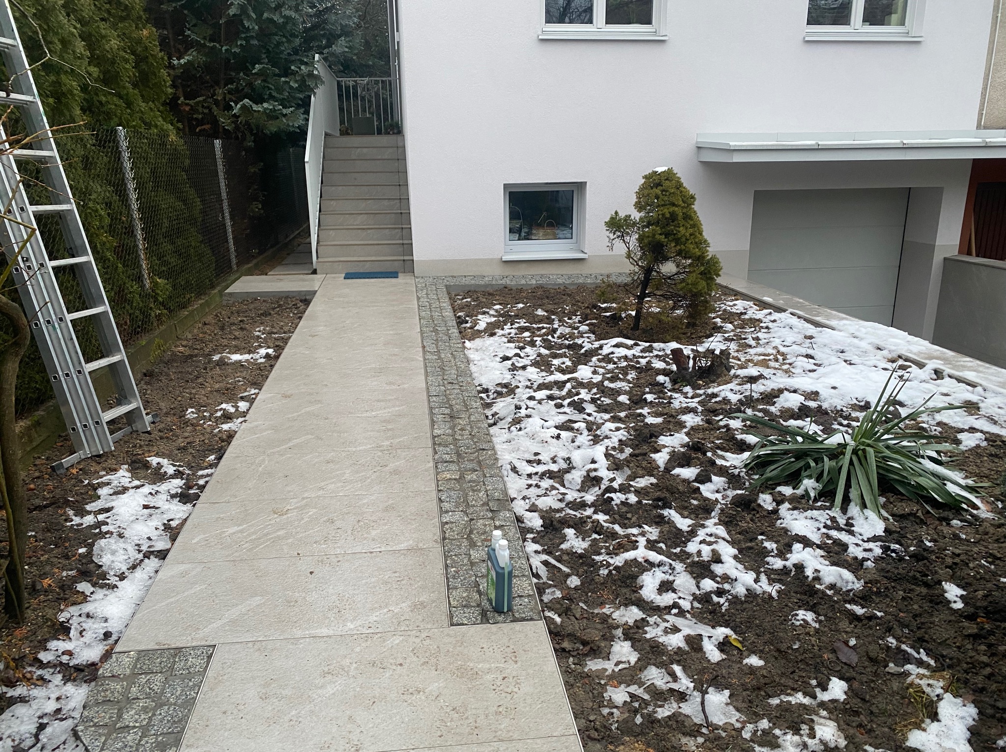 Ułożony chodnik z płyt i kostki brukowej prowadzący do schodów wejściowych domu. Obok rabata z roślinami i resztkami śniegu. Aluminiowa drabina oparta o ogrodzenie.