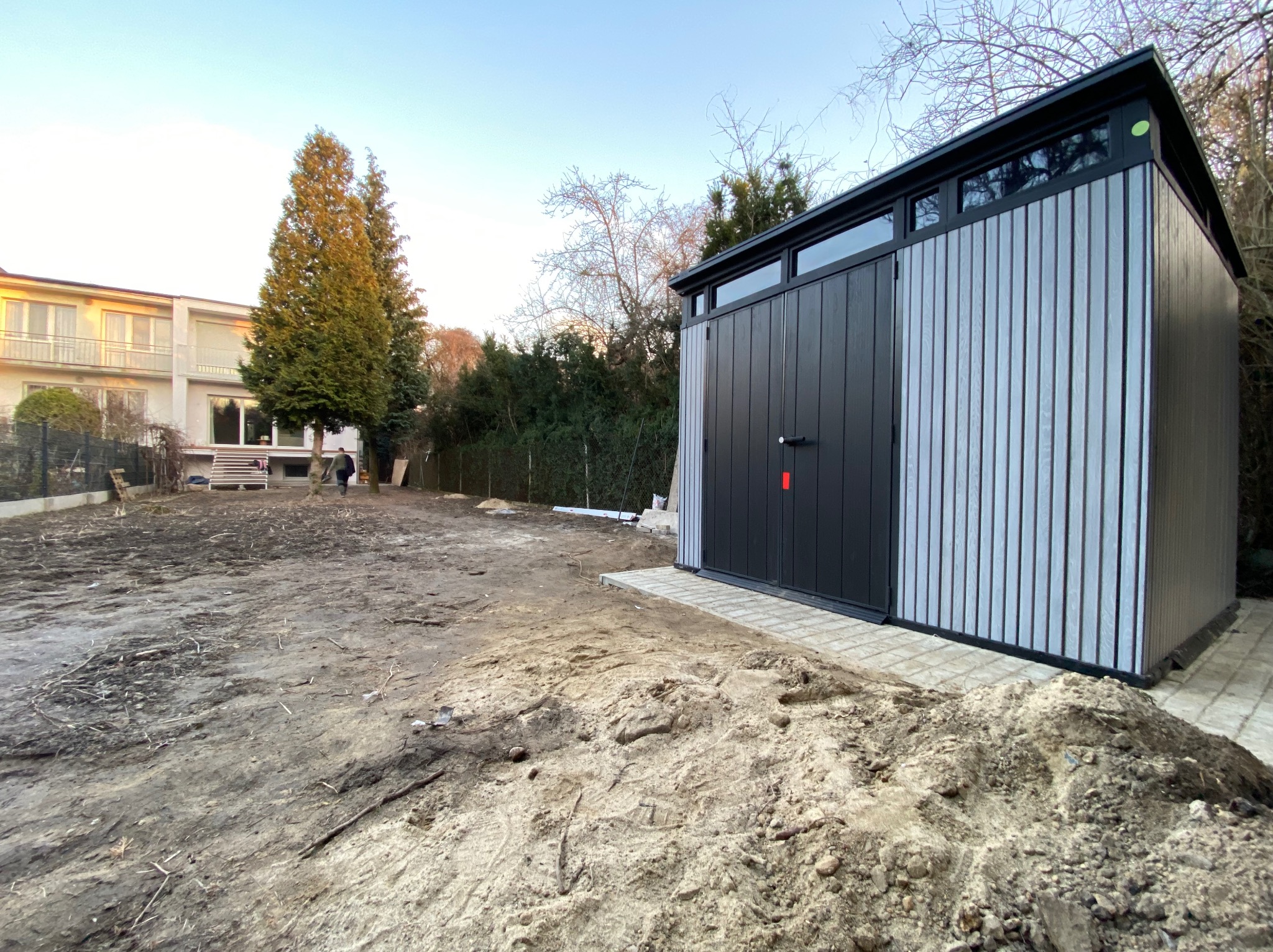 Nowoczesna, grafitowo-szara altana ogrodowa z oknami, ustawiona na kostce brukowej. W tle dom i drzewa, a na pierwszym planie ziemia przygotowana pod trawnik.