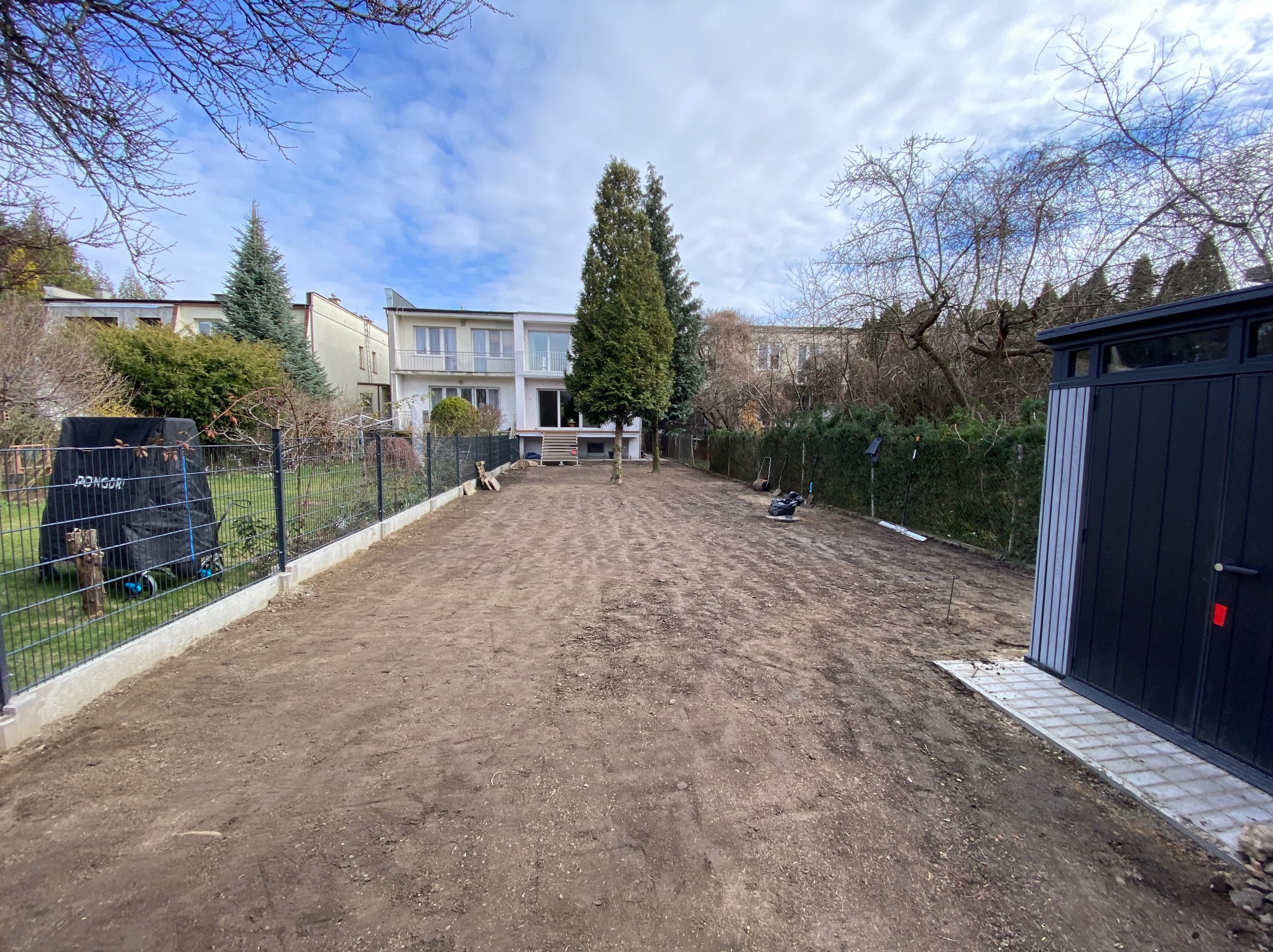 Przygotowany teren pod trawnik z widocznymi śladami po glebogryzarce, ogrodzeniem panelowym i budynkiem w tle, w słoneczny dzień.