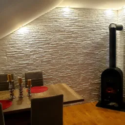 Jadalnia w poddaszu z dekoracyjną ścianą z jasnych płytek imitujących kamień, drewnianym stołem z dekoracjami i czarnym piecem kominkowym.