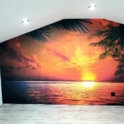 Fototapeta z motywem zachodzącego słońca nad morzem i palmami, umieszczona na ścianie ze skosem w jasnym pomieszczeniu z drewnopodobną podłogą.