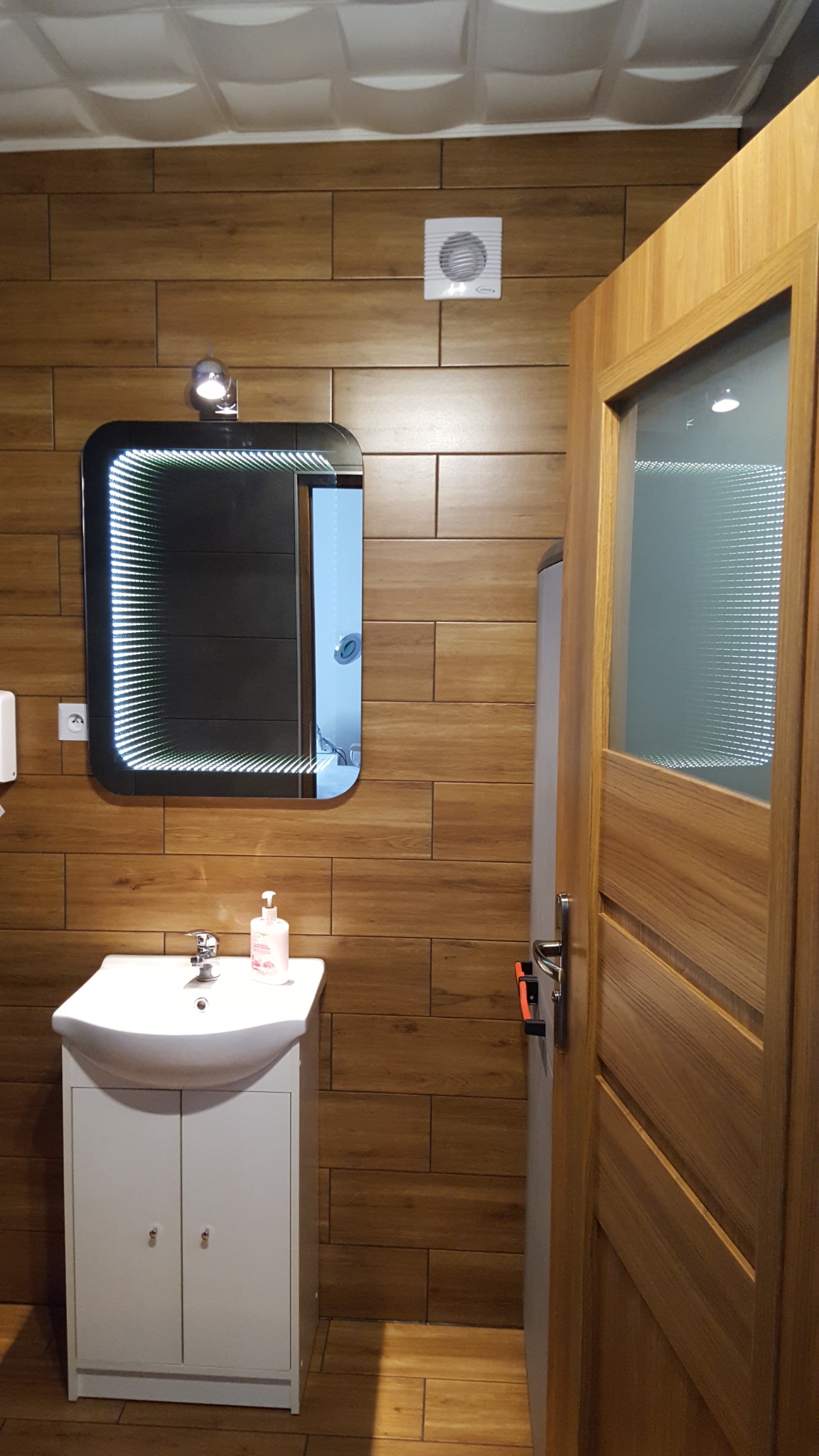 Nowoczesna łazienka z drewnopodobnymi płytkami, podświetlanym lustrem, białą szafką z umywalką i drewnianymi drzwiami z przeszkleniem.