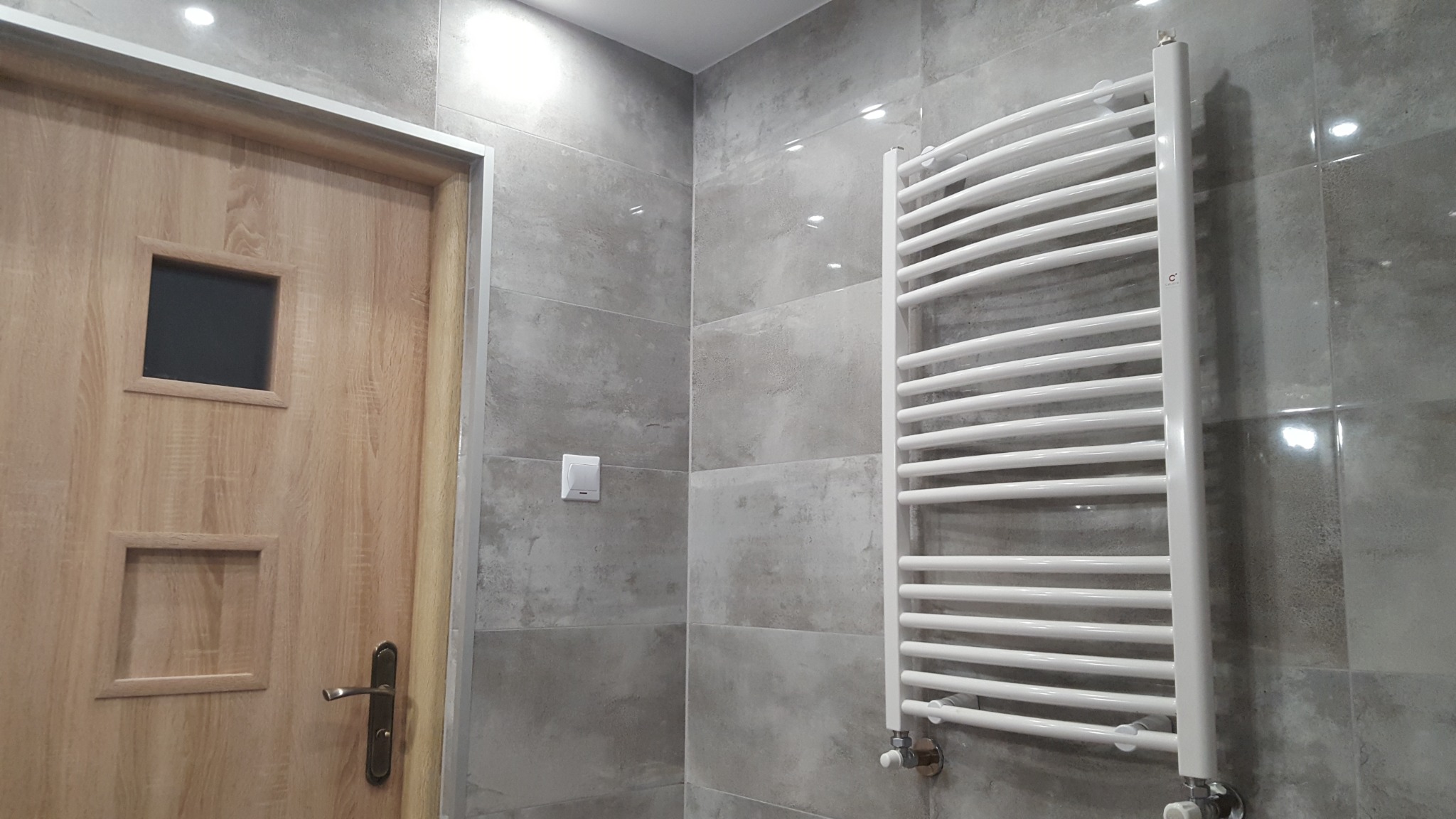 Wykończona łazienka z szarymi, prostokątnymi płytkami, drewnianymi drzwiami z dwoma małymi okienkami i białym grzejnikiem drabinkowym.