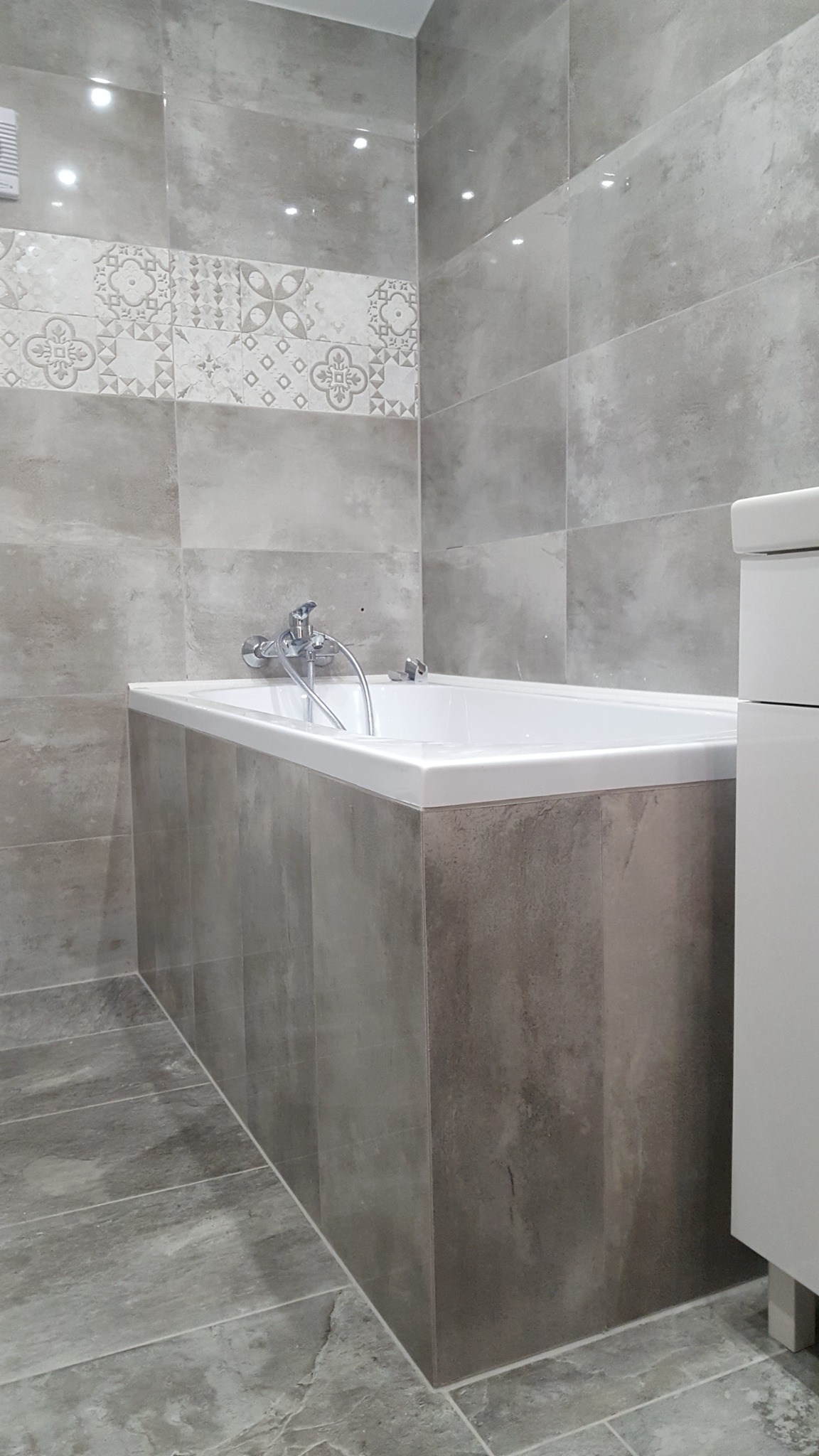 Wykończona łazienka z wanną obudowaną szarymi płytkami, pas płytek dekoracyjnych z motywem kwiatowym, szara podłoga z płytek imitujących kamień, chromowana bateria wannowa.