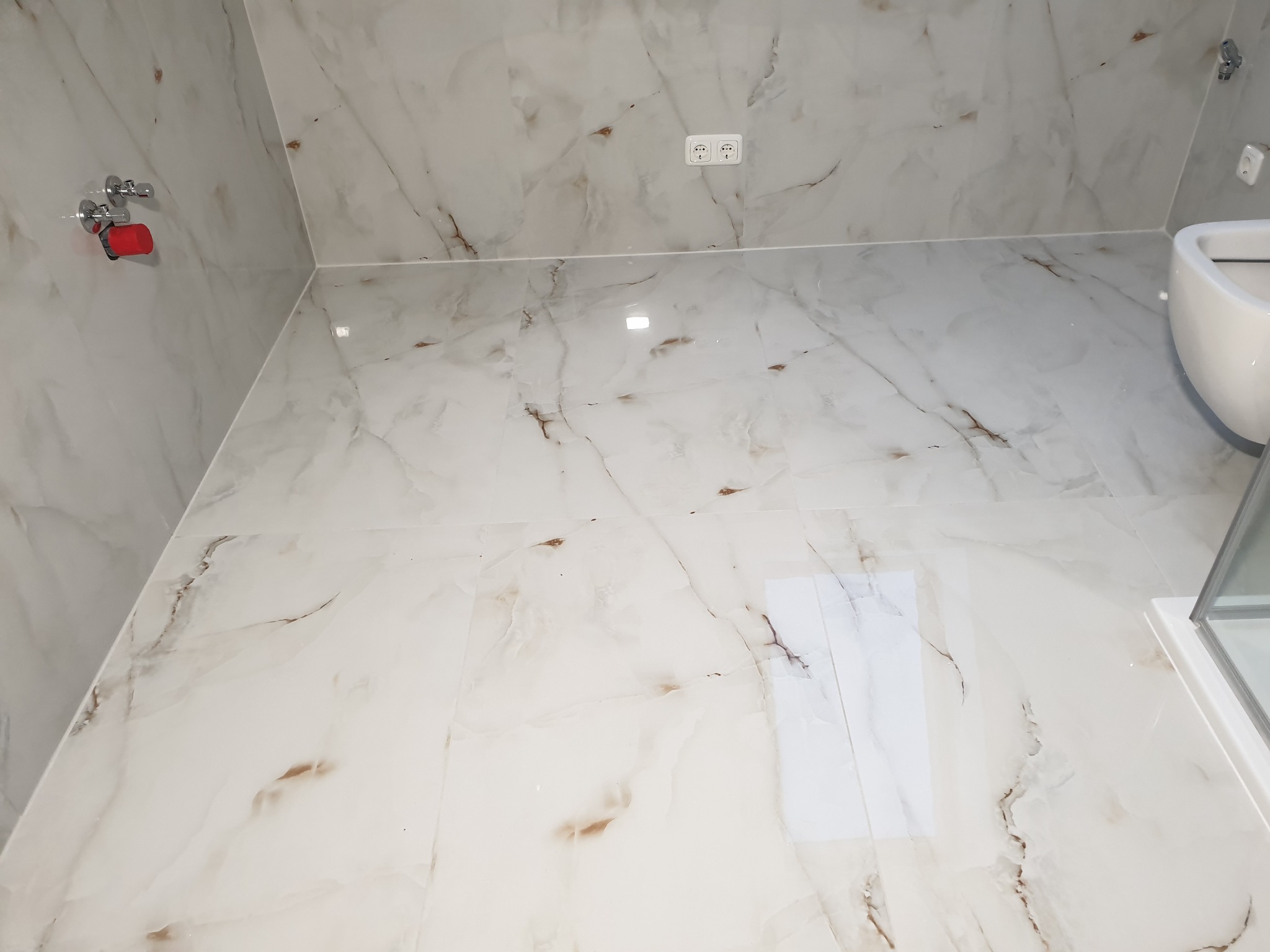 Wykończona łazienka z marmurowymi płytkami na ścianach i podłodze, widoczny fragment umywalki i kabiny prysznicowej, oraz zawór z czerwoną rączką na ścianie.