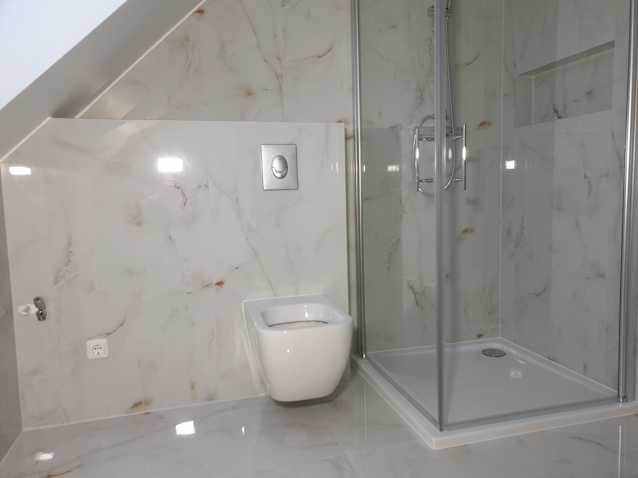 Nowoczesna łazienka na poddaszu z marmurowymi płytkami, wiszącą toaletą z chromowanym przyciskiem spłukiwania i kabiną prysznicową z półką w ścianie.