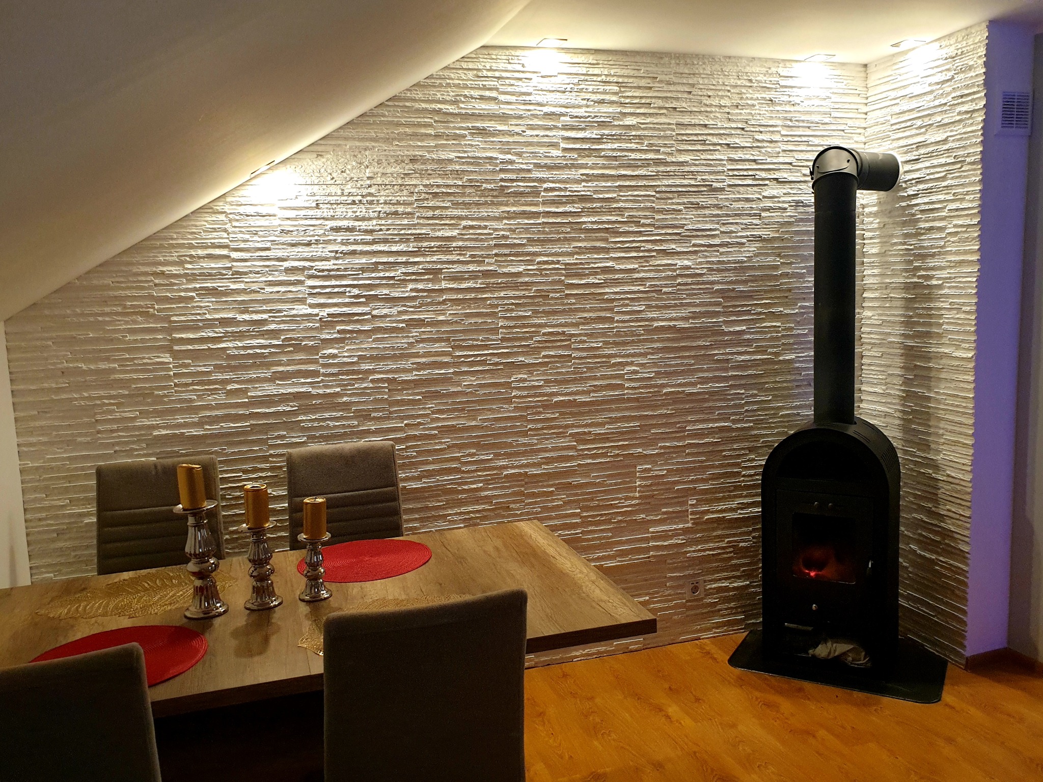 Jadalnia w poddaszu z dekoracyjną ścianą z jasnych płytek imitujących kamień, drewnianym stołem z dekoracjami i czarnym piecem kominkowym.