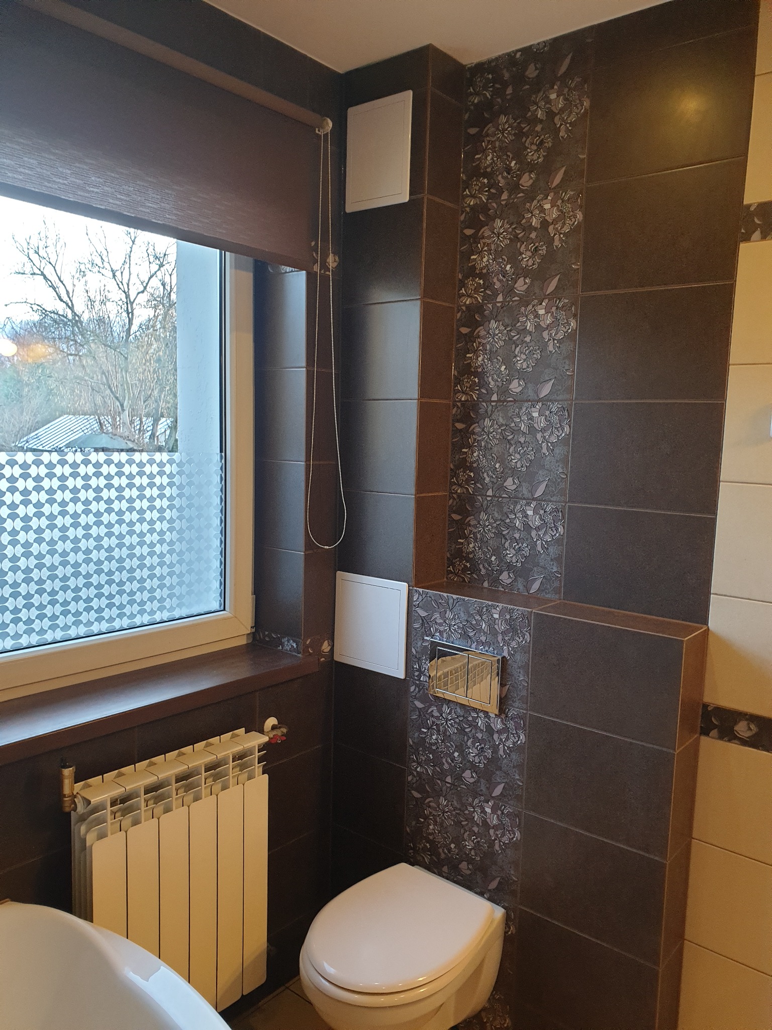 Wykończona łazienka z brązowymi płytkami, dekoracyjnym panelem z motywem kwiatowym, białą toaletą i grzejnikiem pod oknem z roletą.