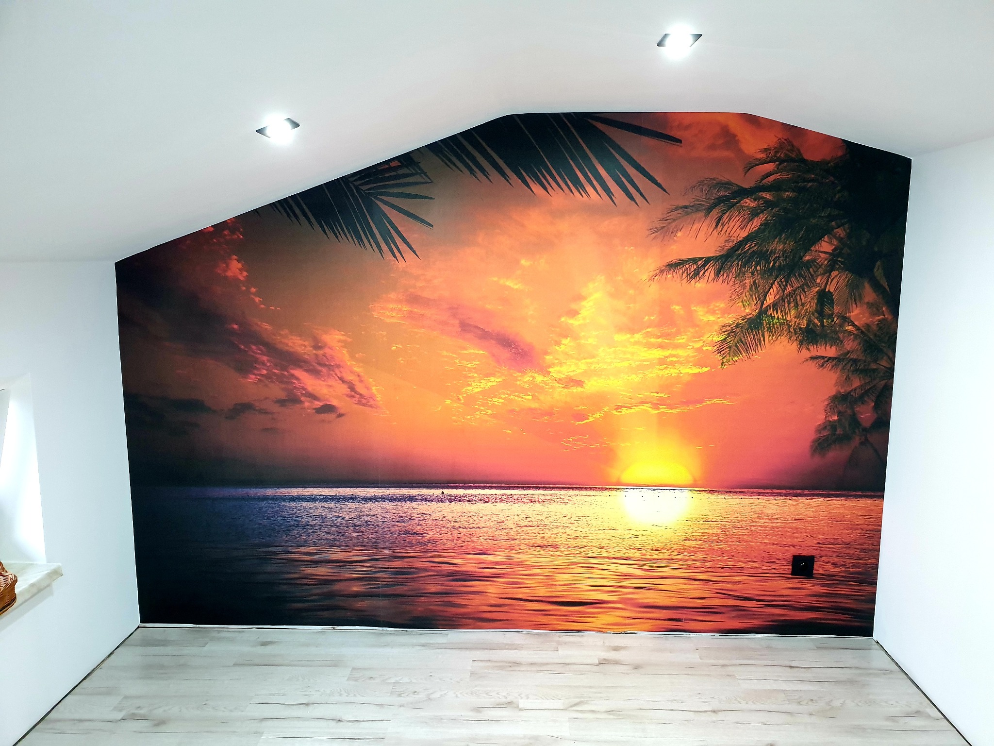 Fototapeta z motywem zachodzącego słońca nad morzem i palmami, umieszczona na ścianie ze skosem w jasnym pomieszczeniu z drewnopodobną podłogą.