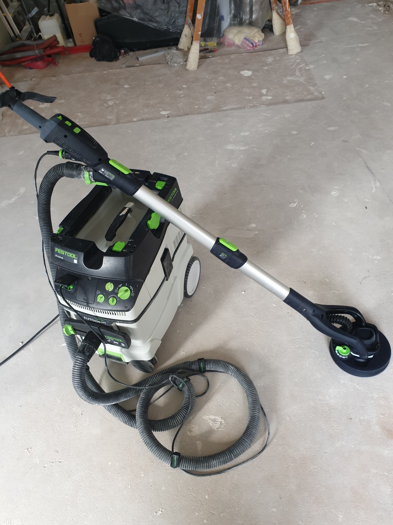 Szlifierka do gipsu marki Festool z podłączonym odkurzaczem przemysłowym, stojąca na surowej, szarej podłodze w trakcie prac wykończeniowych.