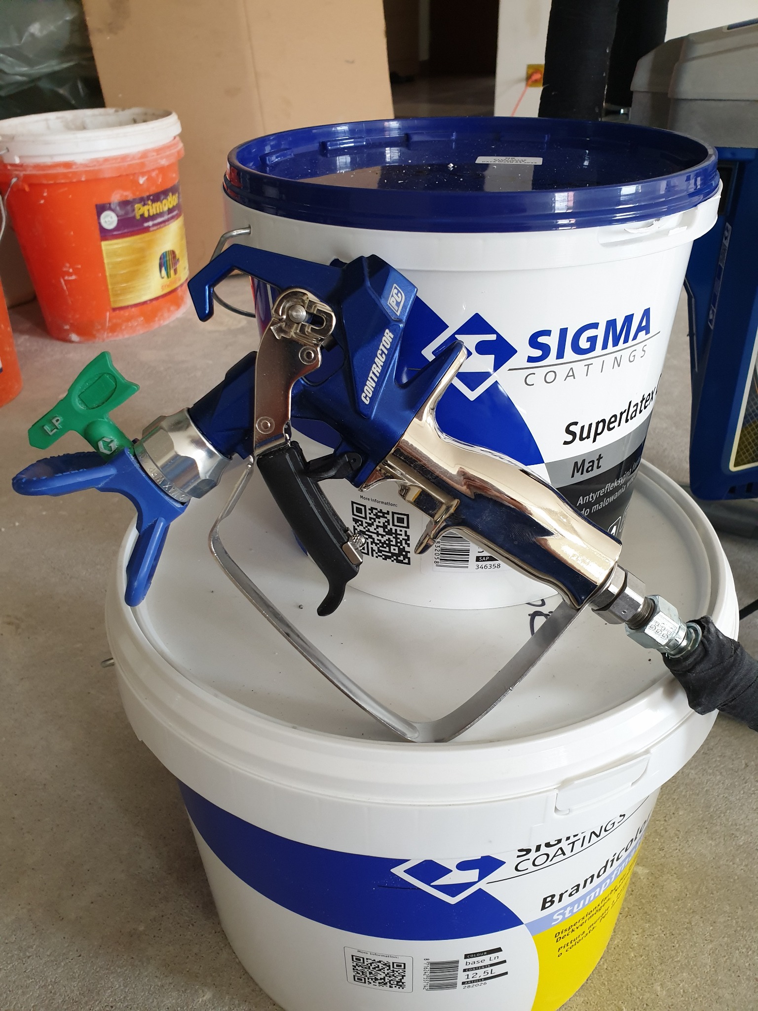 Pistolet natryskowy do farb leżący na wiadrach z farbą Sigma Coatings i Brandicola Stumpfmat, widoczne detale dyszy i węża doprowadzającego.
