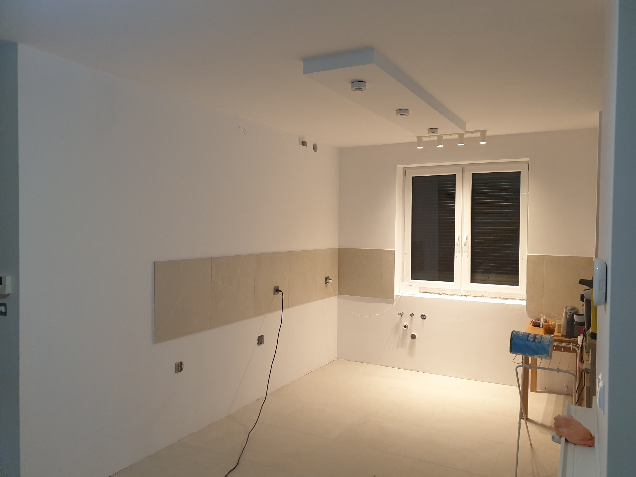 Wnętrze kuchni w trakcie prac wykończeniowych; częściowo położone płytki na ścianie, widoczne punkty instalacji elektrycznej i hydraulicznej, sufit z oświetleniem punktowym.