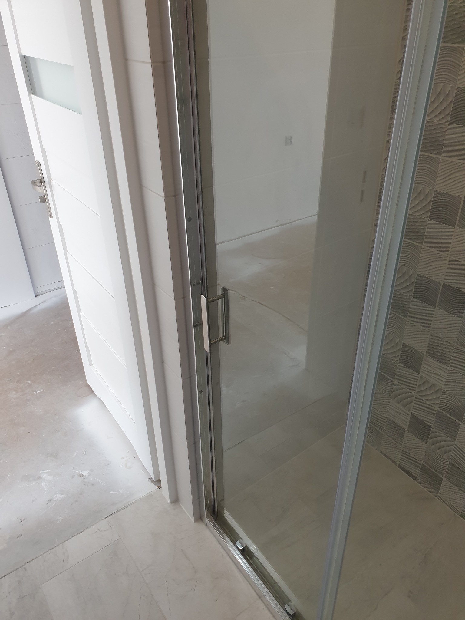 Zamknięta kabina prysznicowa z białymi drzwiami i wzorzystymi płytkami w tle, widoczny fragment podłogi w trakcie prac wykończeniowych.