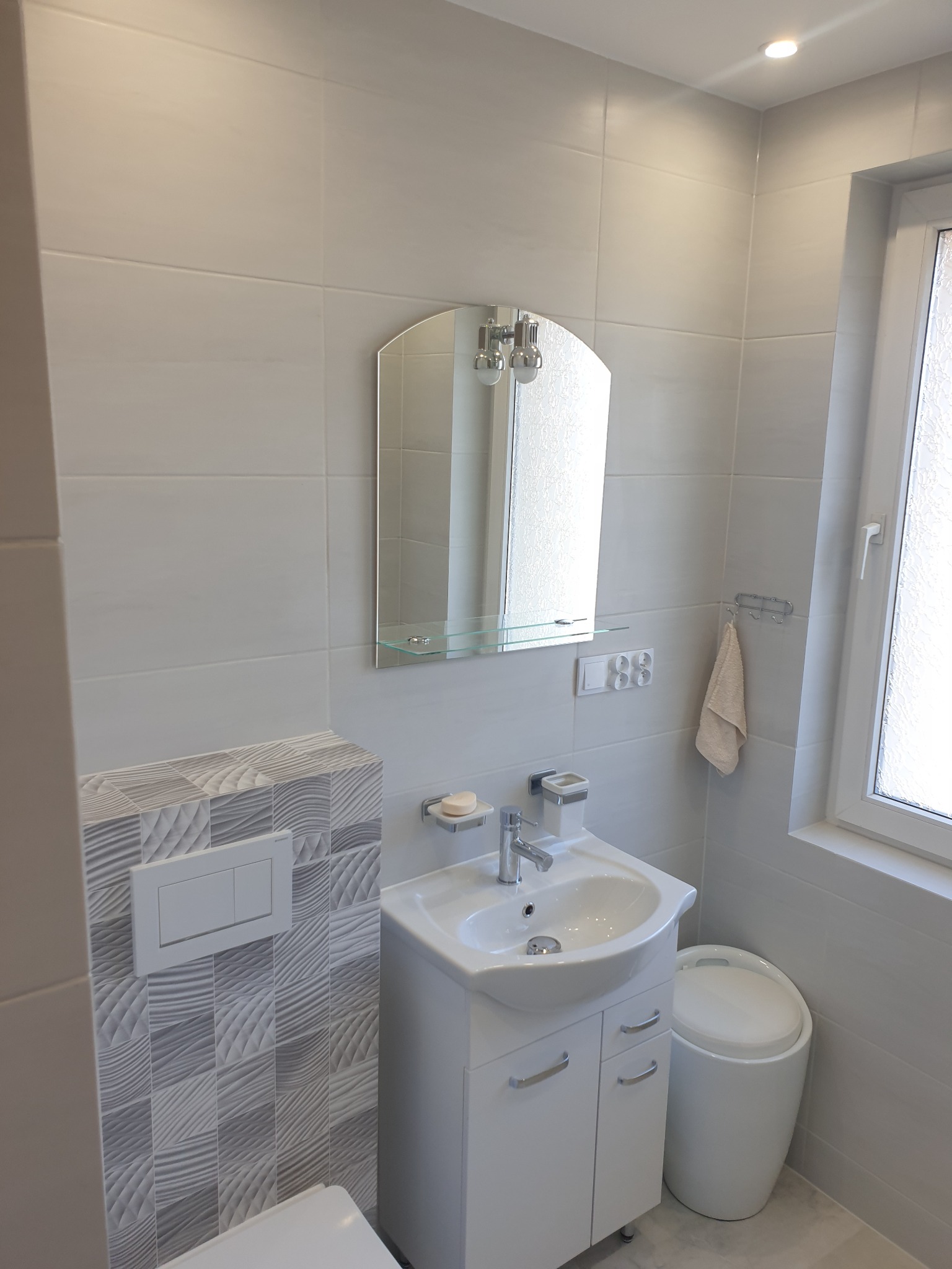 Jasna łazienka po remoncie z umywalką na szafce, lustrem z półką, toaletą z zabudowaną spłuczką i akcesoriami łazienkowymi.