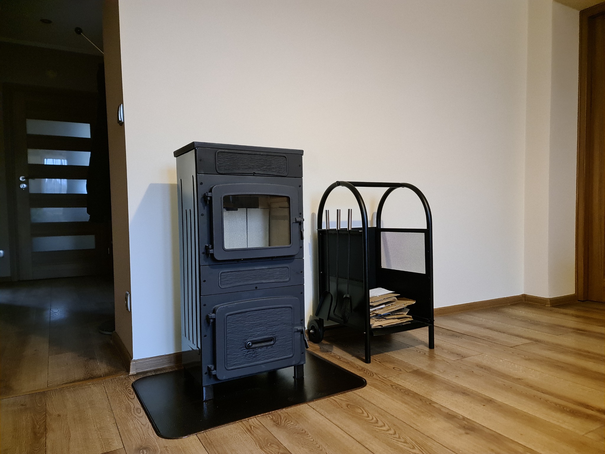 Czarny piec wolnostojący z szybą i metalowym stojakiem na drewno oraz akcesoriami kominkowymi, ustawiony na czarnej macie ochronnej na podłodze z paneli.