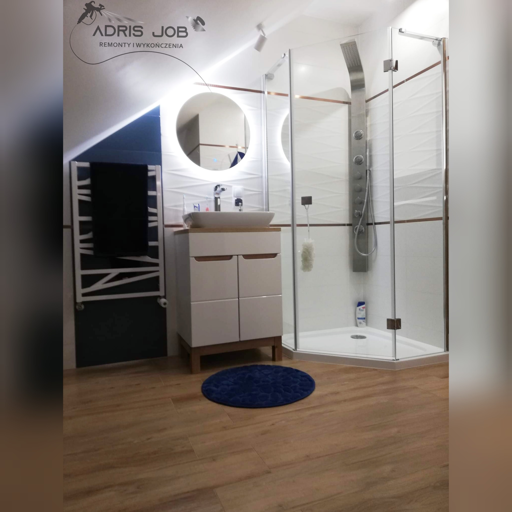 Nowoczesna łazienka na poddaszu z kabiną prysznicową typu walk-in, okrągłym lustrem z podświetleniem LED, białą szafką pod umywalką z drewnianym blatem i grzejnikiem drabinkowym.