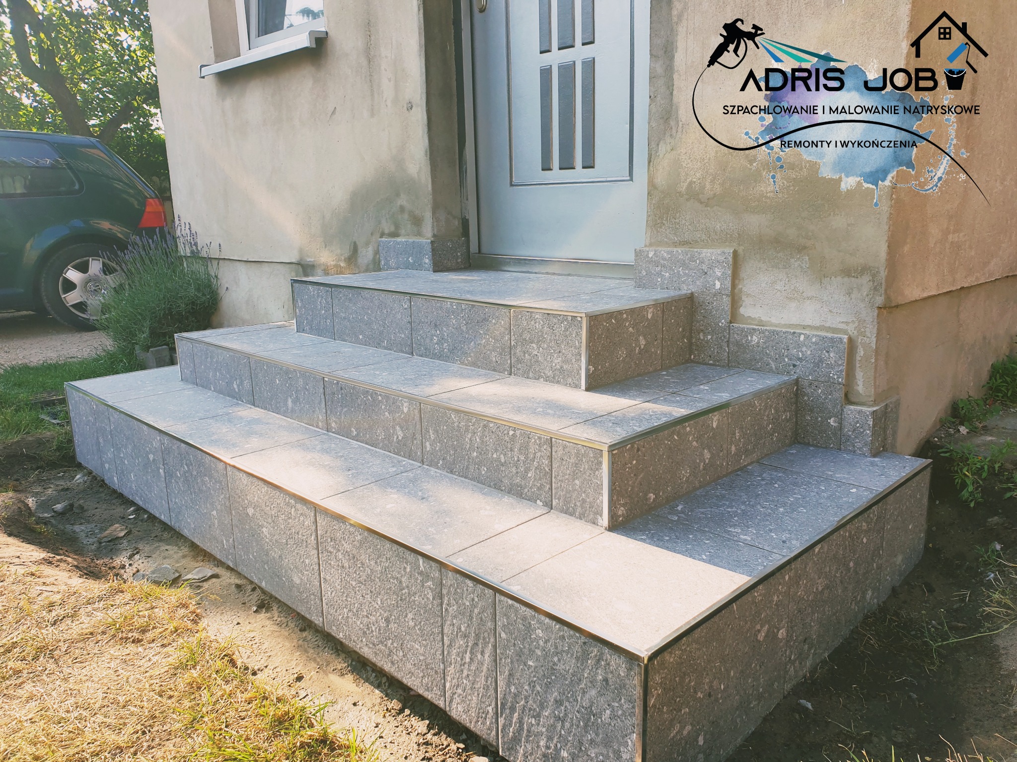 Wykończone płytkami schody wejściowe do domu z szarą elewacją i logo firmy na ścianie.