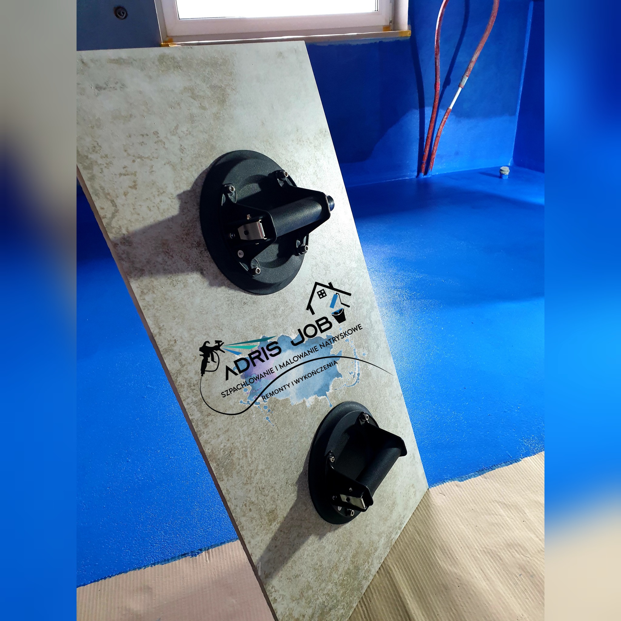Płytka ceramiczna z logo firmy ADRIS JOB przymocowana przyssawkami do pomalowanej na niebiesko ściany w trakcie prac wykończeniowych.