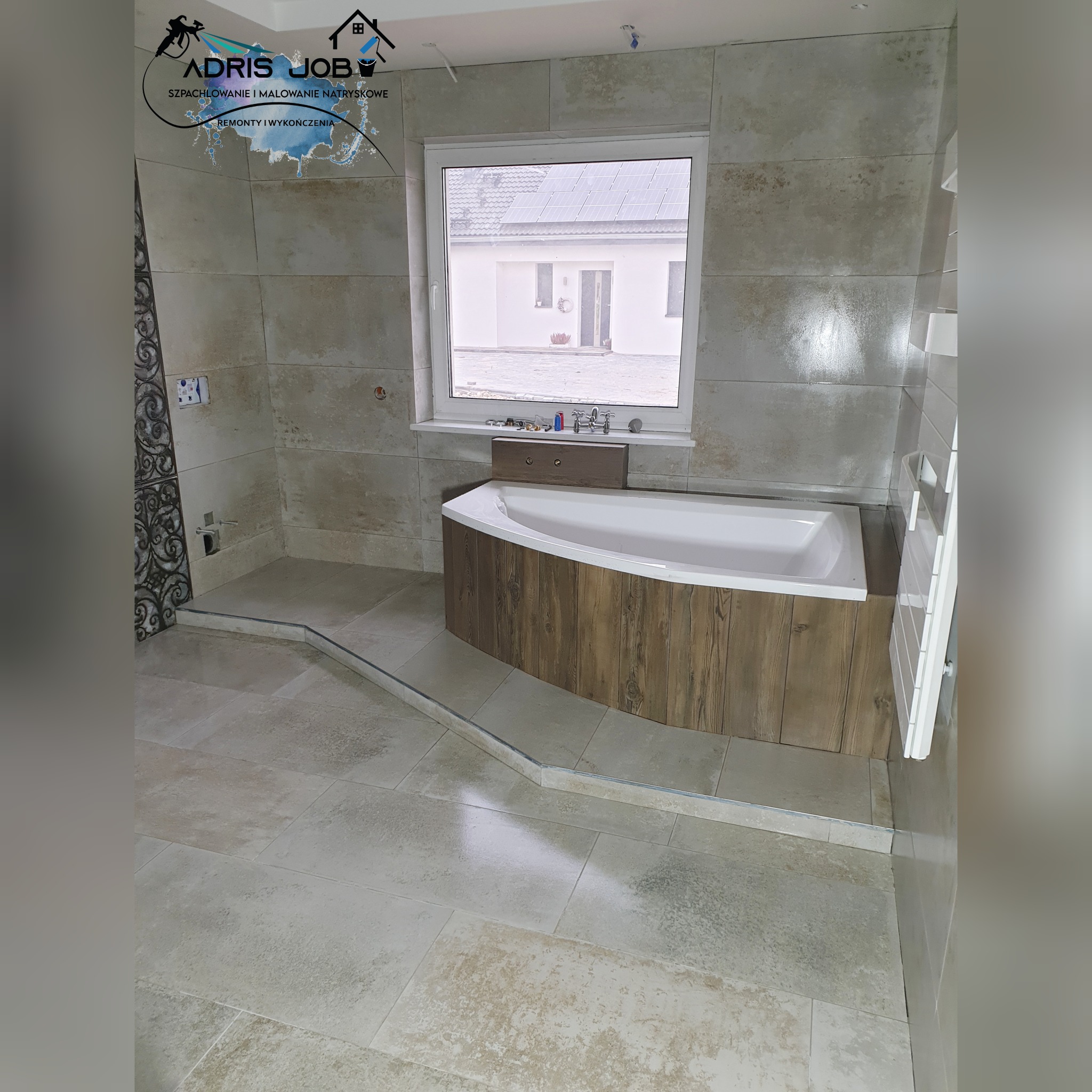 Wykończona łazienka z beżowymi płytkami, obudowana drewnopodobną płytą wanna narożna przy oknie, grzejnik drabinkowy po prawej, logo firmy w lewym górnym rogu.