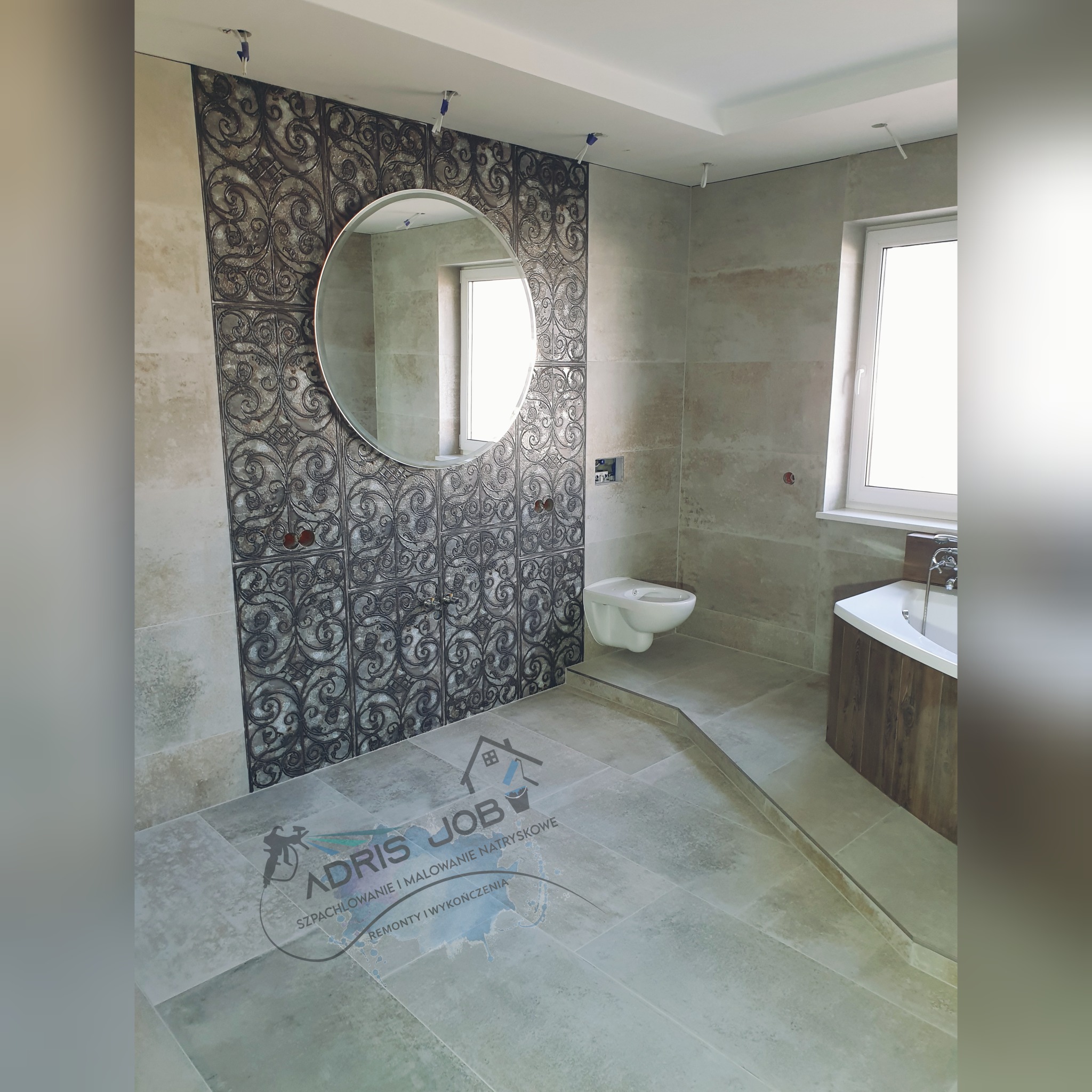 Wykończona łazienka z beżowymi płytkami, dekoracyjną ścianą z wzorem, okrągłym lustrem, podwieszaną toaletą i wanną z drewnianą obudową, widoczny znak wodny firmy na podłodze.