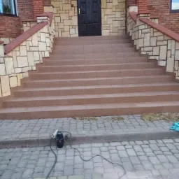 Wykończone schody wejściowe do domu z brązowego kamienia, z widocznym narzędziem elektrycznym na kostce brukowej przed nimi.