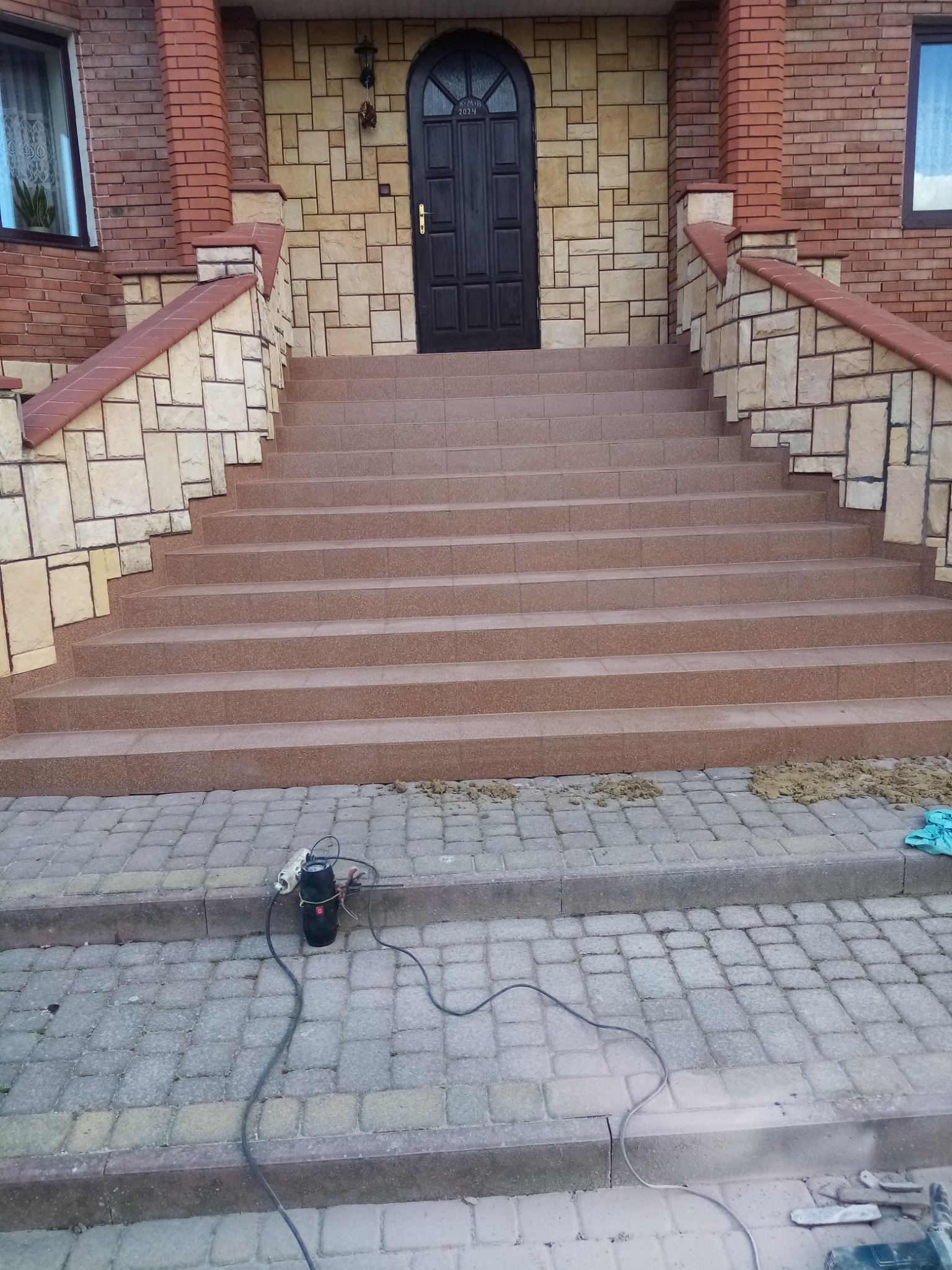 Wykończone schody wejściowe do domu z brązowego kamienia, z widocznym narzędziem elektrycznym na kostce brukowej przed nimi.