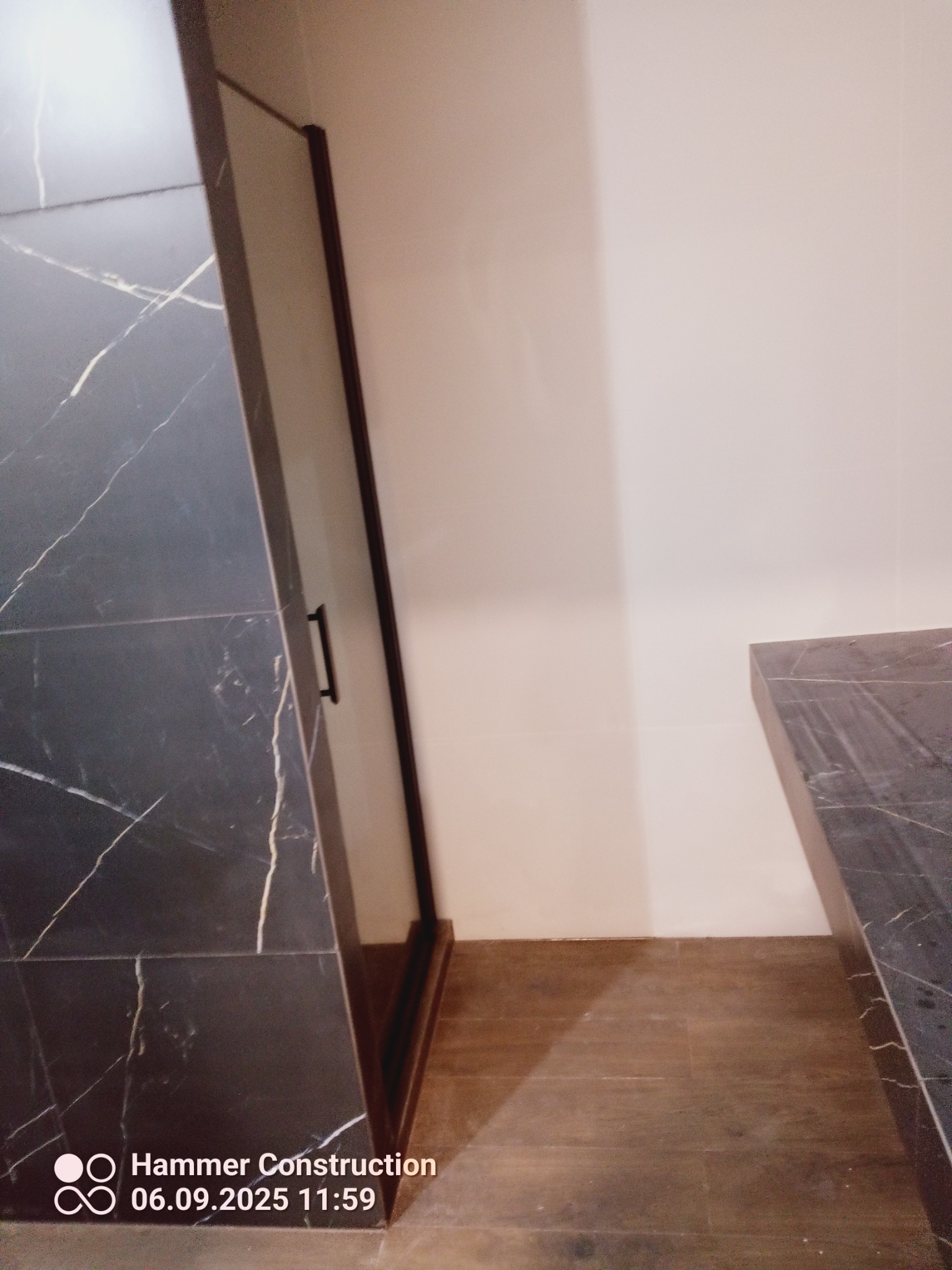 Ciemna kabina prysznicowa z marmurowym wzorem i czarnymi okuciami, widoczna z boku, na tle jasnej ściany i brązowej podłogi.