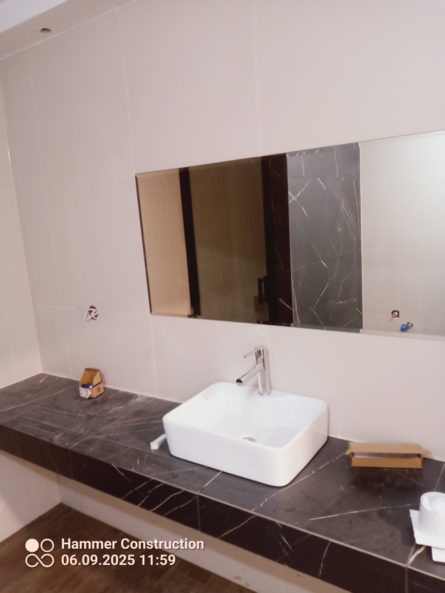 Nowoczesna łazienka z umywalką nablatową i blatem z czarnego marmuru. Widoczne białe płytki i lustro. Umywalka ma chromowaną baterię. Elementy instalacji czekają na podłączenie.