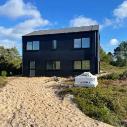 Nowoczesny, dwukondygnacyjny dom z czarną elewacją i szarym dachem na piaszczystym terenie z widocznymi materiałami budowlanymi, otoczony zielenią i drzewami pod błękitnym niebem z chmurami.