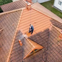 Pracownik w kombinezonie czyści dach z ceglanej dachówki za pomocą myjki ciśnieniowej, widoczne różnice w kolorze czystej i zabrudzonej powierzchni, ujęcie z góry.