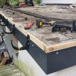 Montaż nowych, czarnych rynien na płaskim dachu z użyciem poziomicy, wkrętarki i liny traserskiej. Dach pokryty papą, widoczne narzędzia i fragment elewacji schodzącej w dół.