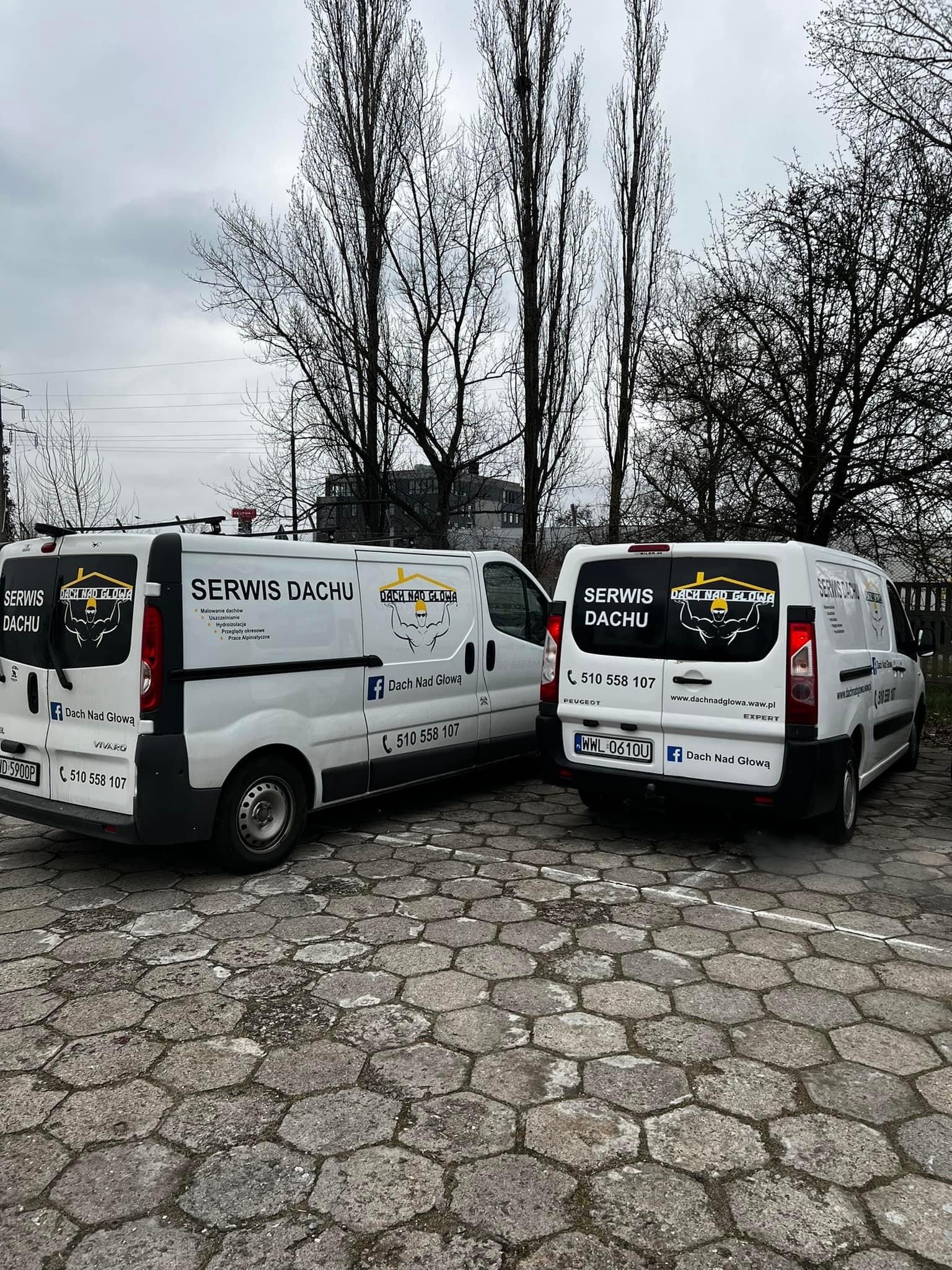 Dwa białe samochody dostawcze z logo firmy dekarskiej 'Dach Nad Głową' zaparkowane na brukowanym parkingu, w tle nagie drzewa i budynki.