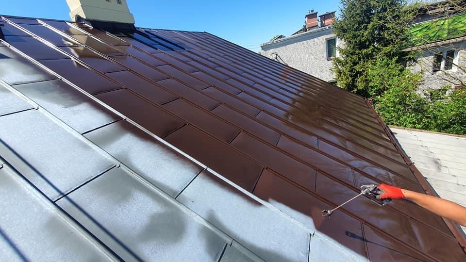 Prace w toku w Warszawie: Częściowo pomalowany na brązowo dach z blachy, z widocznym fragmentem natrysku farby i ręką w czerwonej rękawicy.