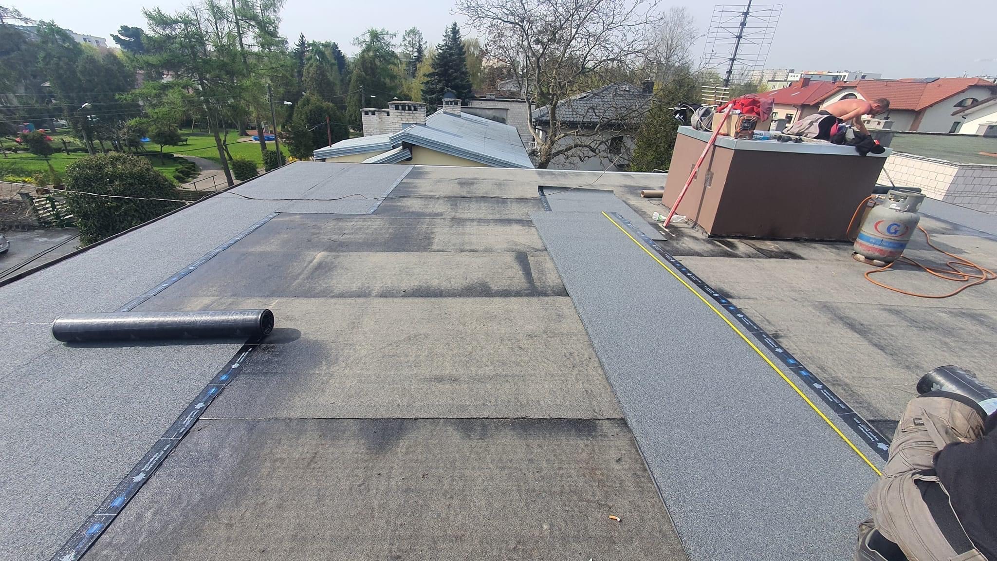 Prace dekarskie na płaskim dachu: częściowo położona papa termozgrzewalna, widoczny cylinder z papą, butla gazowa i dekarz pracujący przy kominie w tle.