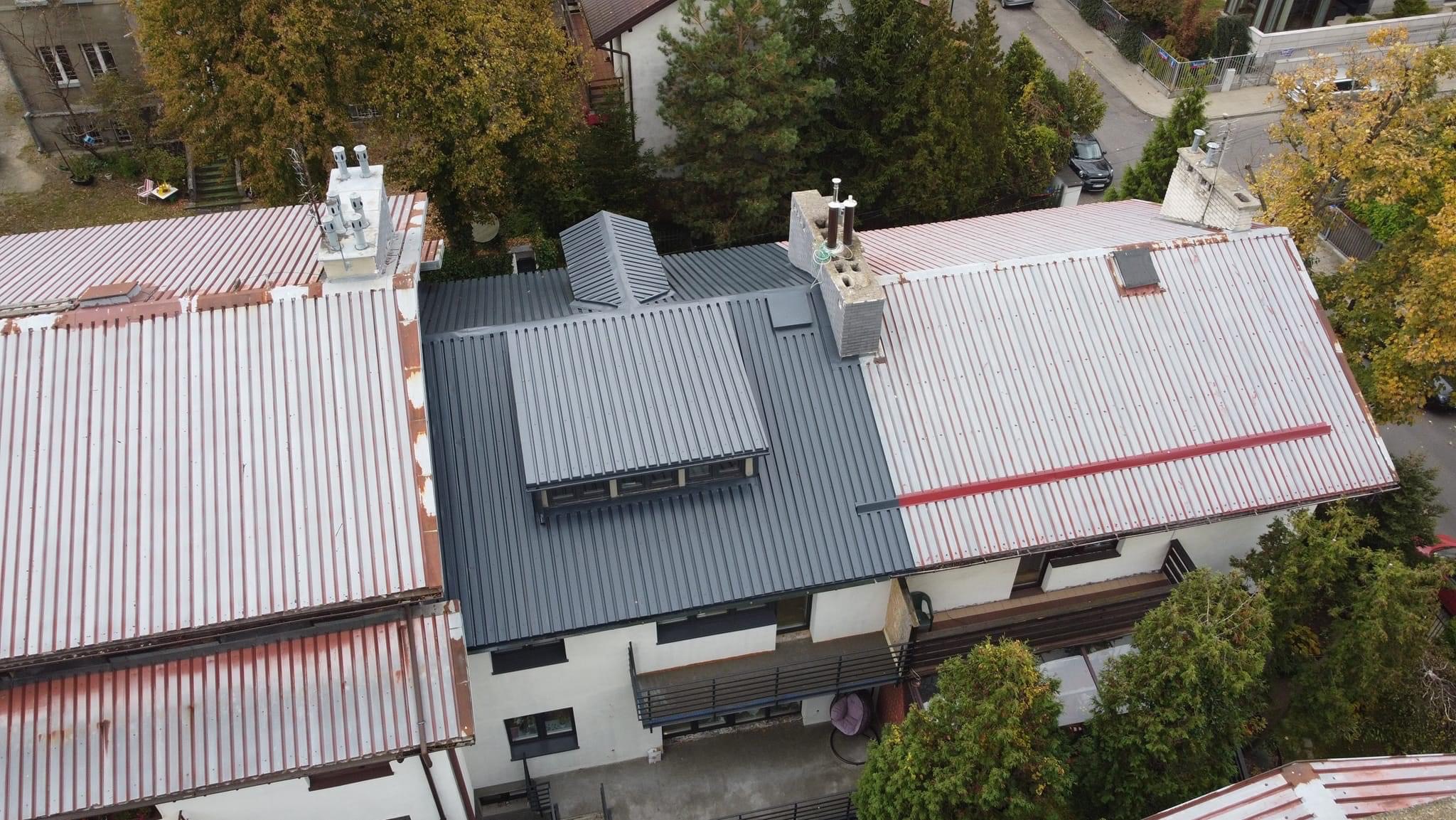 Widok z góry na budynek z częściowo odnowionym dachem z blachy trapezowej w kolorze szarym i białym, z widocznymi kominami i elementami obróbek blacharskich.