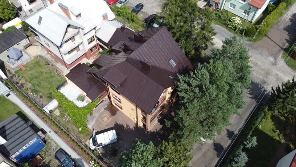 Widok z drona na budynek z ciemnobrązowym, metalowym dachem po renowacji, otoczony zielenią, z zaparkowanym białym vanem na podjeździe.