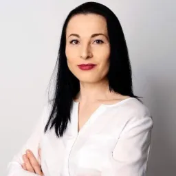 Portret kobiety o ciemnych włosach i czerwonych ustach, ubrana w białą koszulę, na jasnym tle.