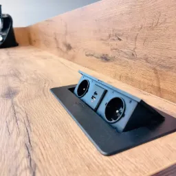 Wpuszczane w blat drewniany, czarne gniazdko elektryczne z portem USB, obok stoi robot kuchenny.