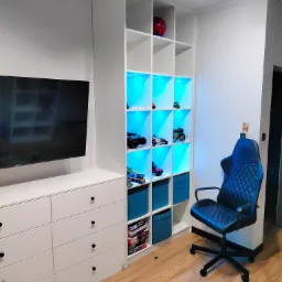 Biała zabudowa meblowa w pokoju dziecięcym z podświetlanymi na niebiesko półkami, na których stoją zabawki i dekoracje, obok biurko i krzesło.