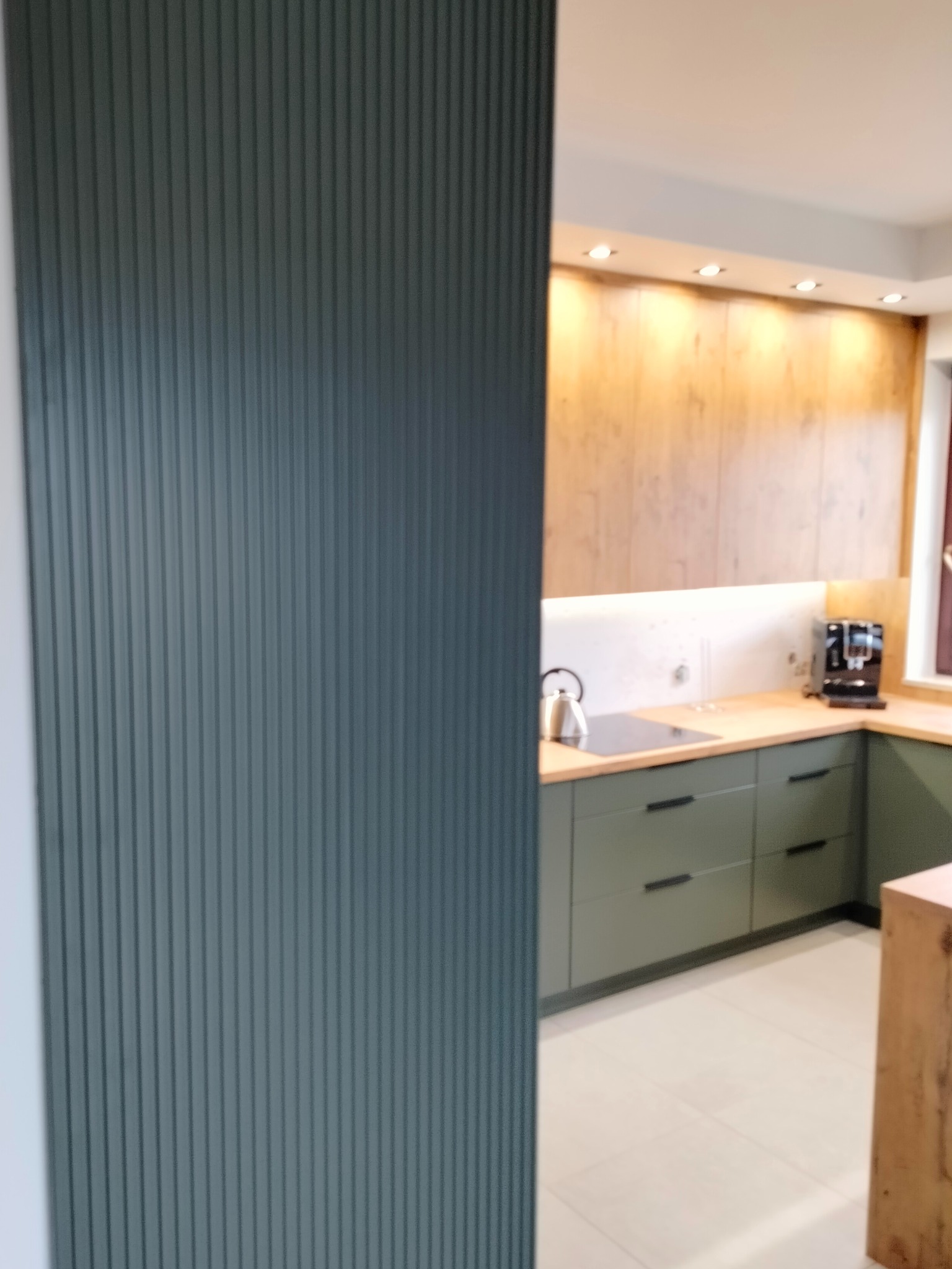 Fragment nowoczesnej kuchni z zabudową w odcieniach zieleni i drewna, widoczny przez szarą, ryflowaną ściankę działową.