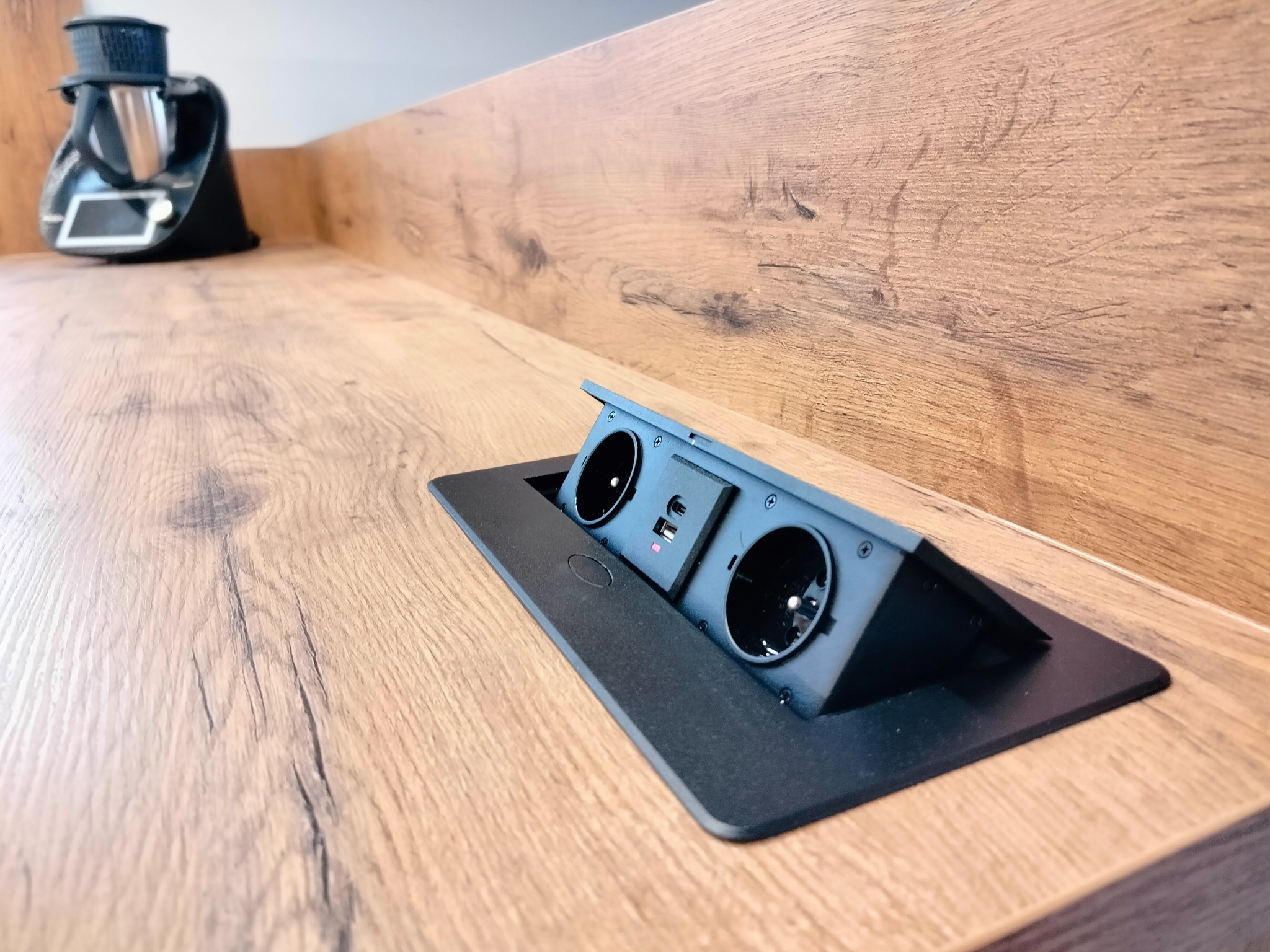 Wpuszczane w blat drewniany, czarne gniazdko elektryczne z portem USB, obok stoi robot kuchenny.