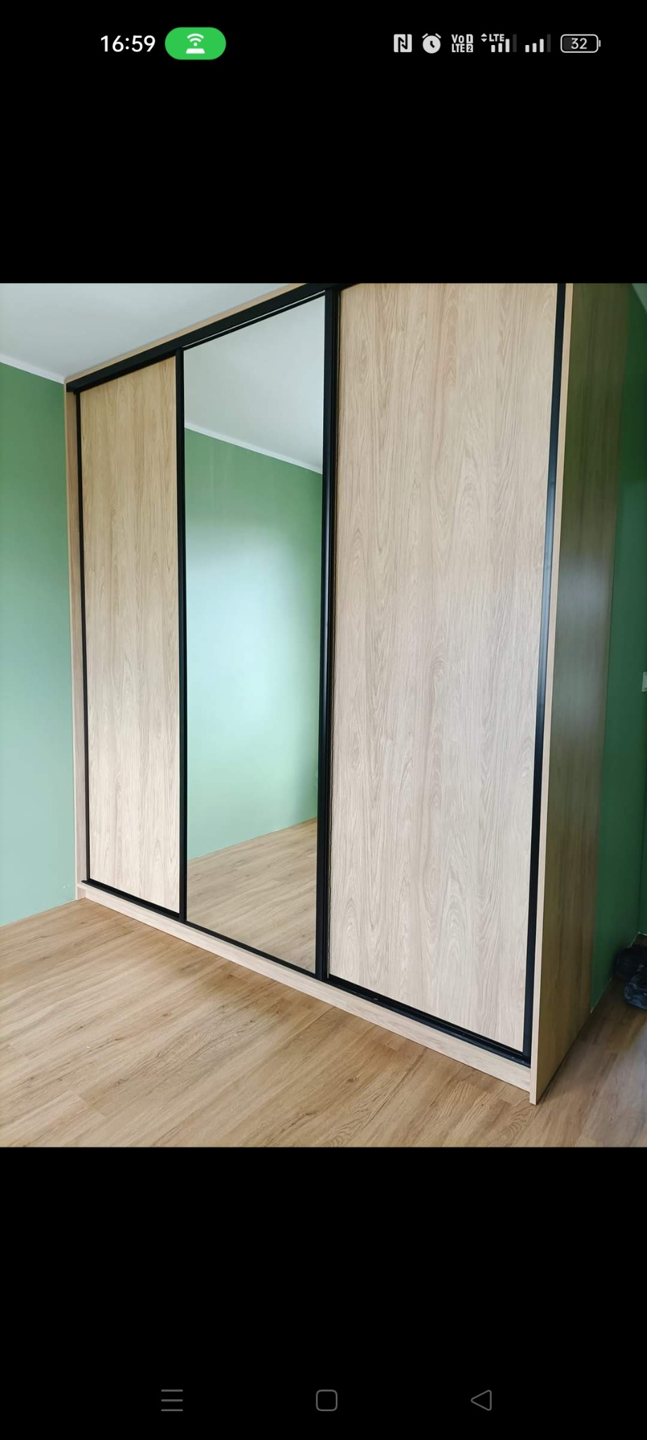 Jasna szafa wnękowa z przesuwnymi drzwiami i lustrem, czarne obramowanie, parkiet na podłodze, miętowa ściana w tle.