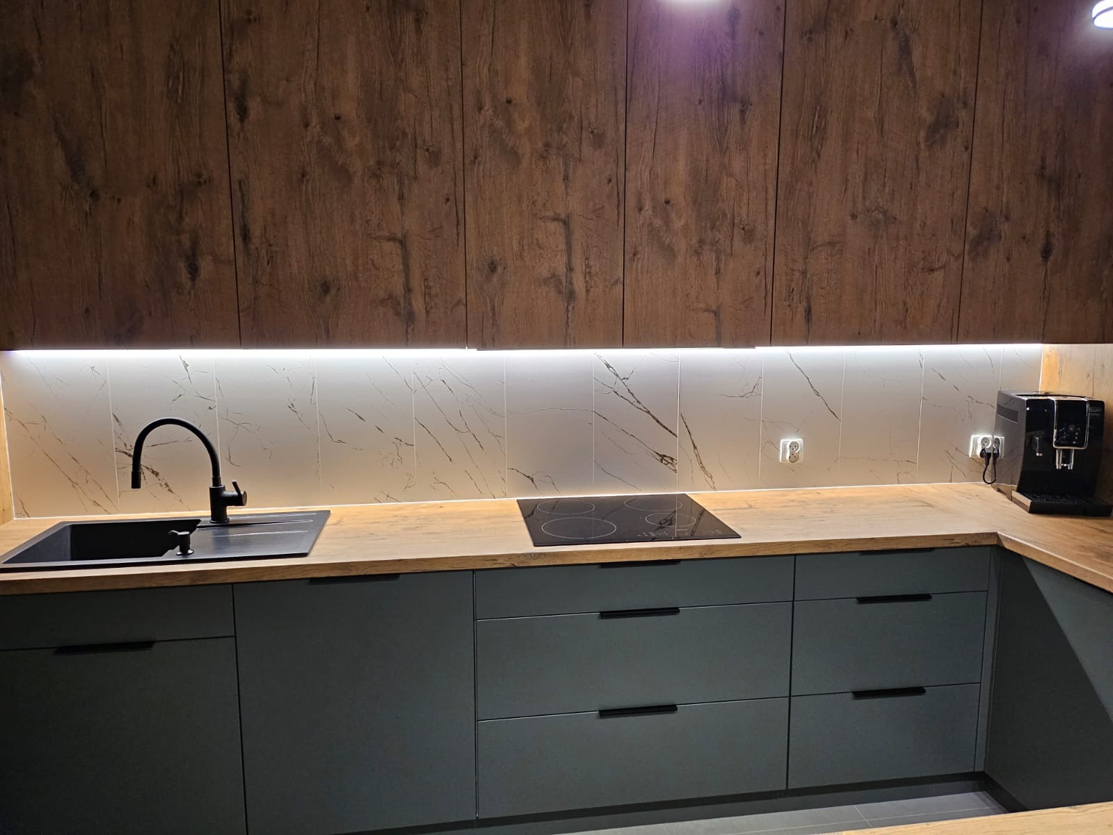 Nowoczesna kuchnia z ciemnymi, matowymi frontami szafek, drewnianym blatem i marmurową ścianą nad blatem. Oświetlenie LED pod szafkami górnymi. Widoczny czarny zlew z baterią i płyta indukcyjna.