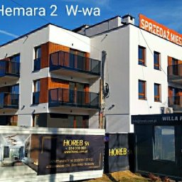 30 lat praktyki - Tomasz Bieliński - Nowoczesny budynek apartamentowy 'Willa Hemara 2' z balkonami z przezroczystymi balustradami, z widoczną reklamą sprzedaży mieszkań na dachu i banerami reklamowymi firmy deweloperskiej Horeb SA...