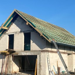 30 lat praktyki - Tomasz Bieliński - Budowa domu jednorodzinnego w trakcie realizacji: murowane ściany z betonu komórkowego, drewniana konstrukcja dachu pokryta zieloną membraną, drabina oparta o budynek, pracownik na dachu.