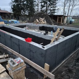 30 lat praktyki - Tomasz Bieliński - Betonowe fundamenty domu z izolacją pionową, widoczny fragment muru z bloczków betonowych, drewniane szalunki i narzędzia budowlane w otoczeniu działki.