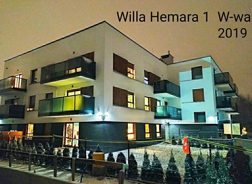 Nocny widok na nowoczesny, trzypiętrowy budynek mieszkalny z balkonami, oświetlony z zewnątrz, z widocznymi choinkami przed budynkiem i napisem 'Willa Hemara 1 W-wa 2019' na górze.